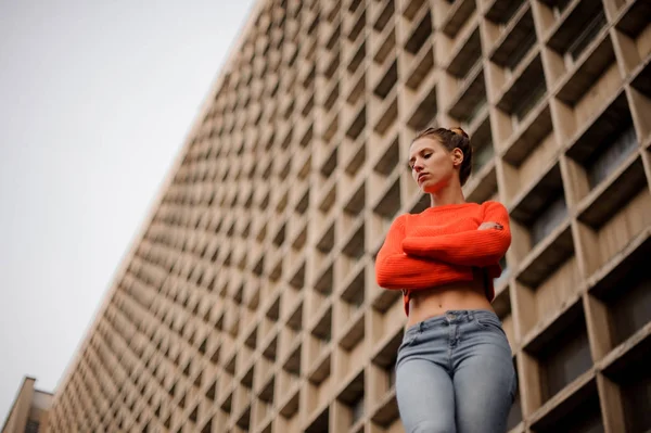 Žena v oranžovém svetru a džínách, stojící na pozadí betonové stavby — Stock fotografie