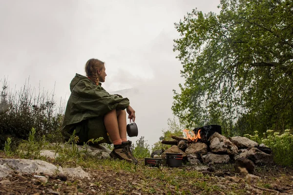 Dziewczynka w płaszczu przeciwdeszczowej siedzi w pobliżu garnka na ognisku — Zdjęcie stockowe