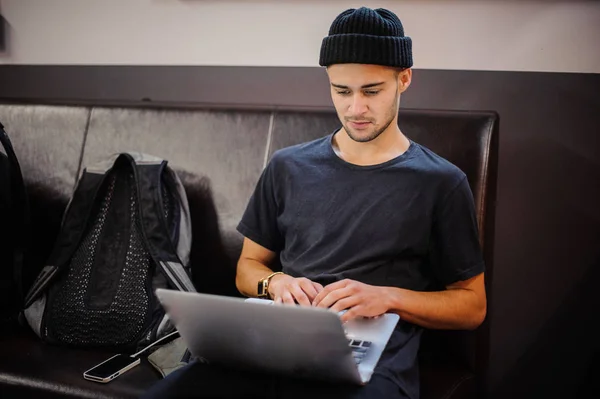 Jonge hipster man in zwarte hoed zittend op de lederen bank en het gebruik van laptop — Stockfoto