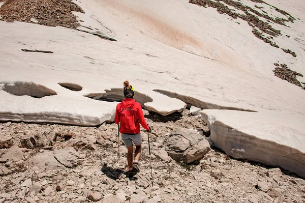 Mujer vista trasera caminando sobre la nieve sucia y rocas con bastones de senderismo — Foto de Stock