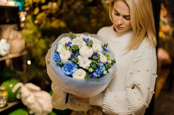 Mujer joven sosteniendo un ramo con flores azules y blancas envueltas en papel transparente mate decorado con hojas verdes —  Fotos de Stock