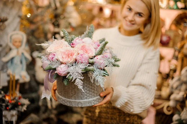 Mosolygó nő kezében egy szürke pulóver minta doboz világos rózsaszín bazsarózsa rózsák díszített fenyőfa ágak — Stock Fotó