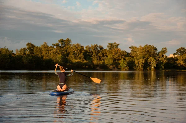 Vista posteriore della donna che siede su tavola di sup tiene pagaia e galleggia sul fiume — Foto Stock