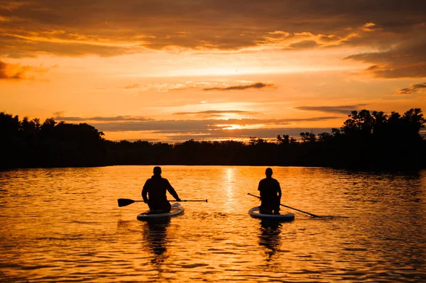 Vista traseira de duas pessoas sentadas em pranchas de jantar que flutuam no rio ao pôr do sol — Fotografia de Stock