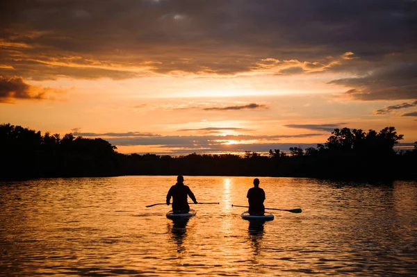 Hermosa vista trasera de dos personas sentadas en tablas de sup que flotan en el río al atardecer —  Fotos de Stock