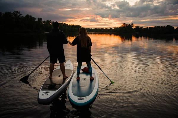 Uomo e donna in piedi su tavole di sup e si tengono per mano al tramonto — Foto Stock