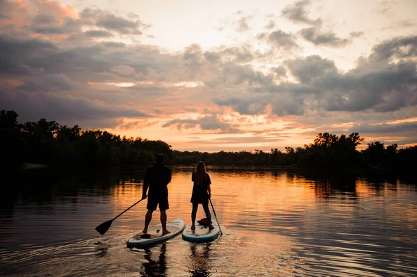 Vista posteriore su uomo e donna in piedi su tavole di sup e galleggianti al tramonto — Foto Stock