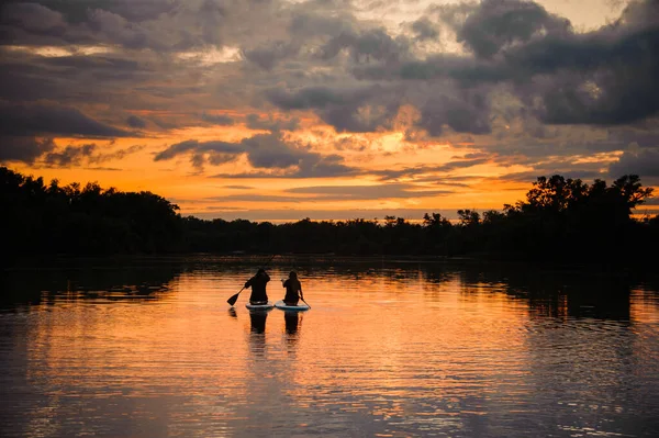 Gran vista del horizonte y la puesta de sol y el río en el que un par de personas nadan en las tablas de sup —  Fotos de Stock