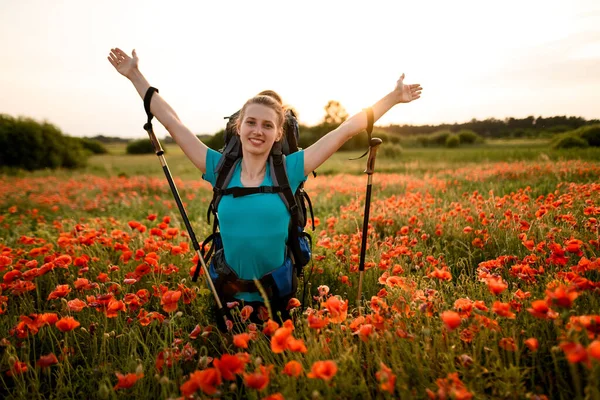 Young smiling woman with backpack stands on poppy field with hands up — Stock Photo, Image