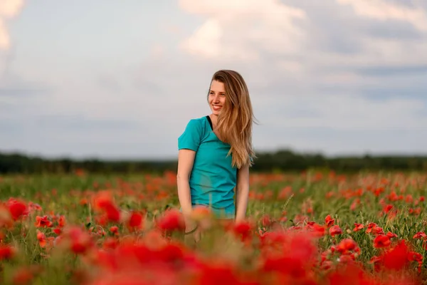 Jovem sorrindo mulher loira com cabelos longos em pé no campo com papoilas vermelhas . — Fotografia de Stock