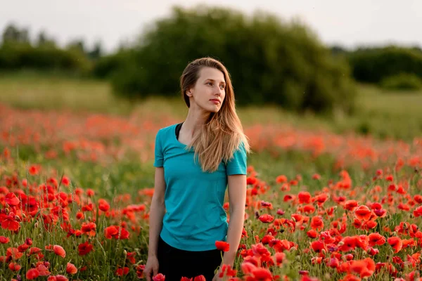 Bela jovem loira mulher com cabelos longos em pé no campo com papoilas vermelhas e olha para o lado . — Fotografia de Stock