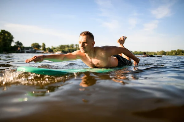 Mann liegt auf Surfbrett, rudert mit den Händen und schwimmt auf dem Fluss. — Stockfoto