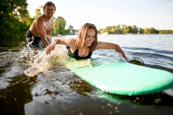 Visa på glad kvinna som ligger på surfbrädet och ror sina händer och manliga instruktör stöder henne — Stockfoto