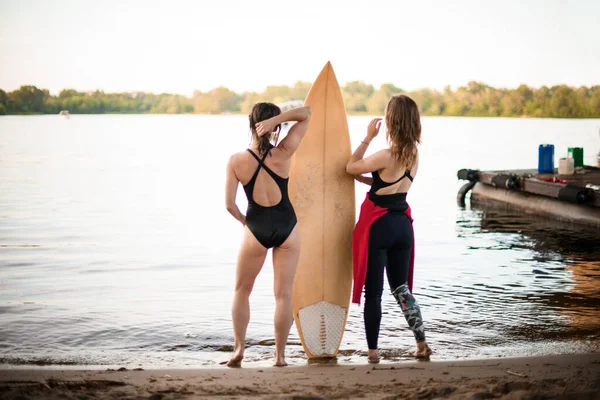 Visszapillantás két fiatal nőre, akik a folyóparton állnak és szörfdeszkát tartanak — Stock Fotó
