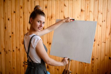 Gülümseyen kadın sanatçı sehpanın yanında duruyor ve tuvali tutuyor.