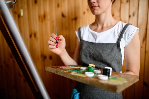 Vrouw kunstenaar houdt palet met multi-gekleurde verf en borstel en tekent op doek — Stockfoto