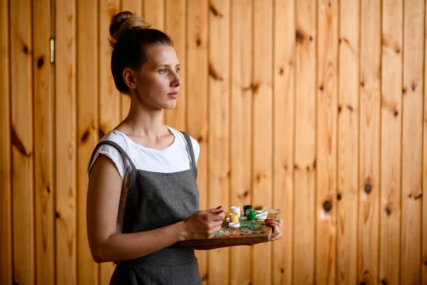 woman with palette and brush in her hand and stands on wooden wall background and looks away