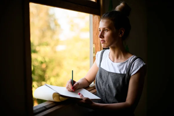 Belle fille avec bloc-notes en papier se tient près de la fenêtre et regarde dehors. — Photo