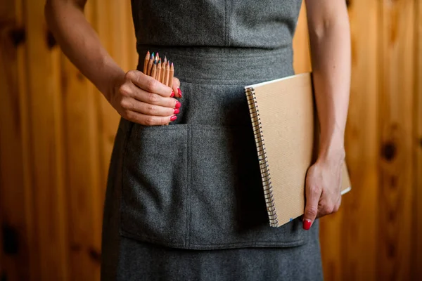 Женщина в сером фартуке держит в руках пачку цветных карандашей и альбомов — стоковое фото