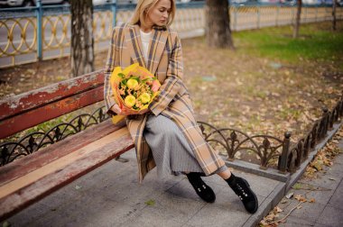 Ekose ceketli kadın elinde sarı bir buket çiçekle bankta oturuyor..