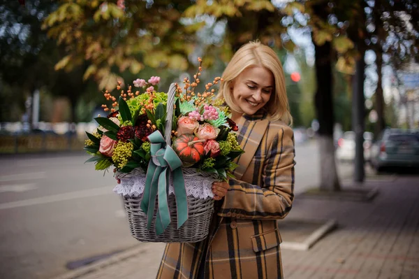 Χαρούμενη ξανθιά γυναίκα στο παλτό κρατά καλάθι με λουλούδια στο εσωτερικό. — Φωτογραφία Αρχείου