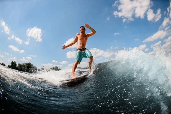 Cheerful athletic guy actively ride on the waves on surfboard against blue sky — Stock Photo, Image