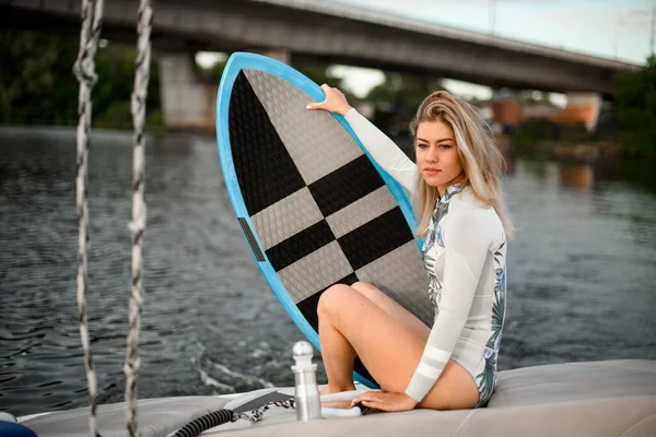 young attractive woman in swimsuit sits on boat and holds surfboard with her hand