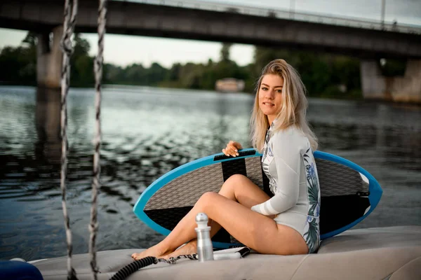 Jeune belle femme en maillot de bain s'assoit sur le bateau et tient la planche de surf avec sa main — Photo