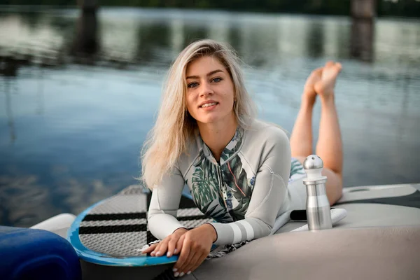 Mooie blonde vrouw in grijs zwempak ligt op boot met surfplank in de buurt. — Stockfoto