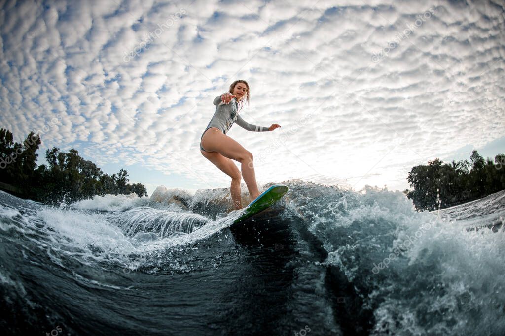 young beautiful blonde woman rides wave on surfboard against cloudy sky background