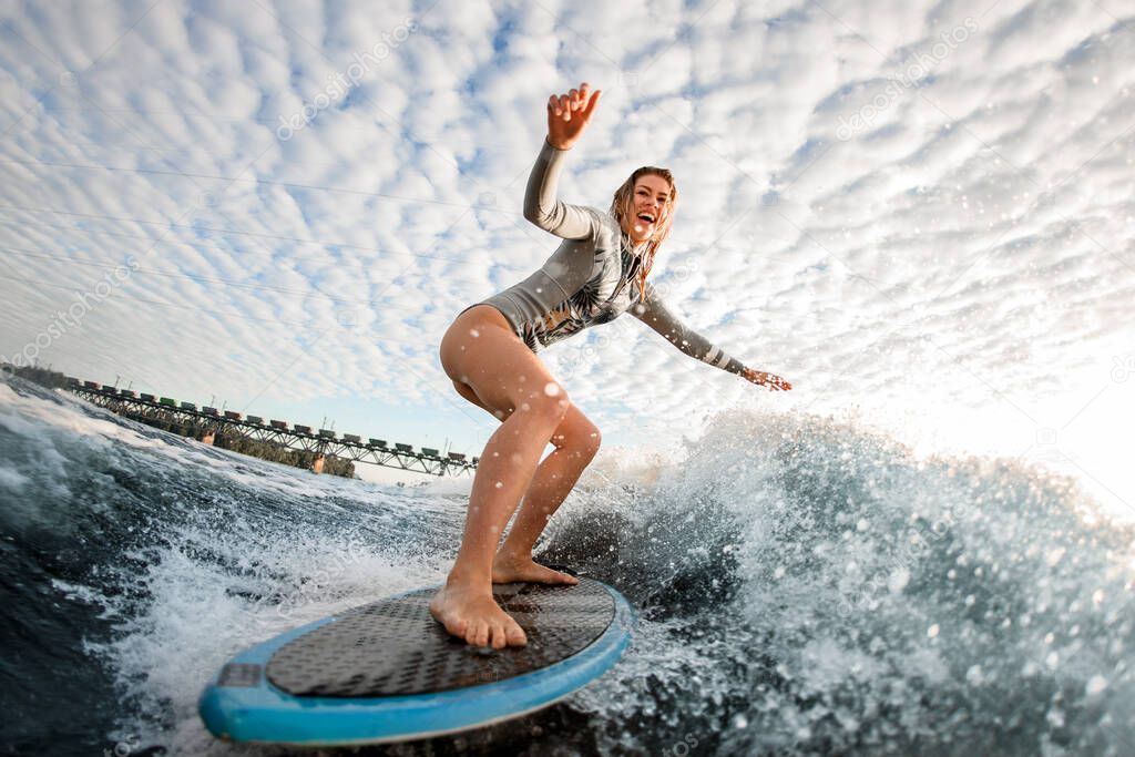 cheerful blonde woman rides wave on surfboard against cloudy sky background