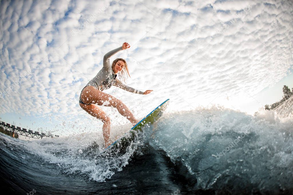 young blonde woman rides up on wave on surfboard against cloudy sky background
