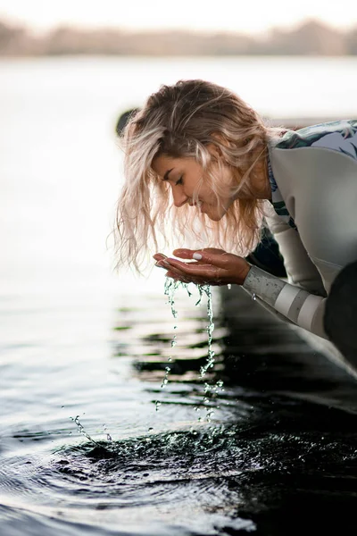Vrouw met blond haar verzamelt water met haar handen uit de rivier. — Stockfoto