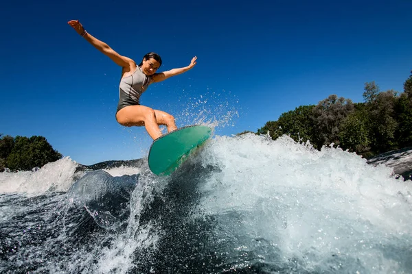 Mujer feliz wakesurfer en traje de baño gris salta con tabla de surf brillante —  Fotos de Stock