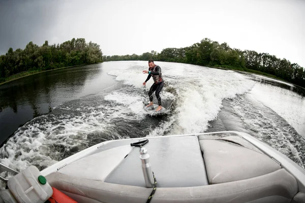 若い男はボートの後ろの波の上にサーフボードに乗る. — ストック写真