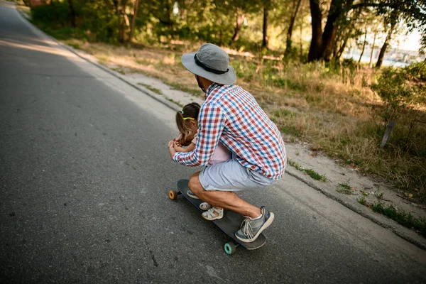 Visão traseira no homem que, juntamente com sua filha andando de skate na estrada no parque de verão — Fotografia de Stock