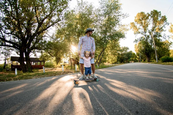 Homem adulto está montando skate com sua filhinha na estrada no parque — Fotografia de Stock