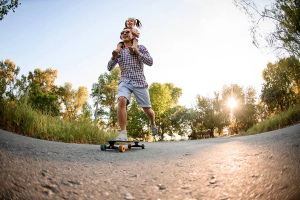 Jovem alegre segura menina em seus ombros e andar de skate no parque — Fotografia de Stock
