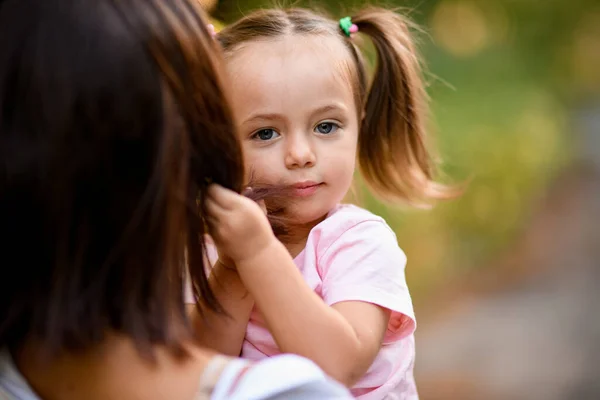 Retrato de niña que se sienta en los brazos de su madre y juega con el pelo — Foto de Stock