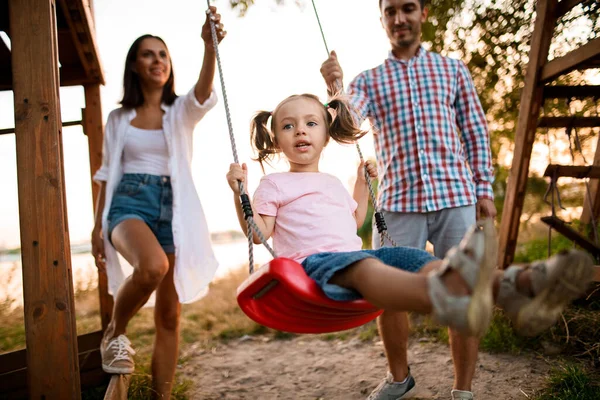 Cheerful young father and mother swing their cute little daughter on swing — Stock Photo, Image