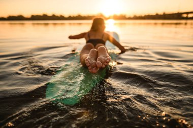 Wakeboard 'da uzanan ve arka planda yüzen bir kadının kıyı şeridi ve sabah güneşi.