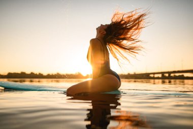 Sörf tahtasına oturmuş, güneşin doğuşunu seyreden uzun saçlı bir kadın.