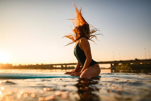 Вид сбоку привлекательной женщины с длинными струящимися волосами, сидящей на доске для серфинга на фоне заката солнца — стоковое фото