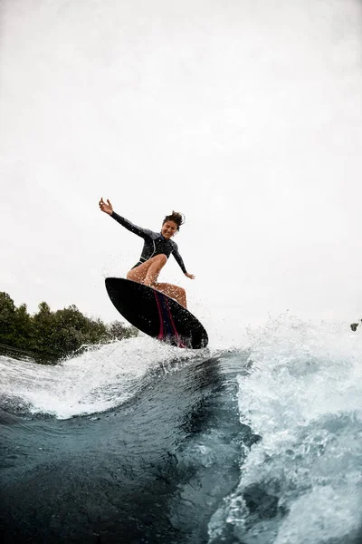 Fröhliche Frau im schwarzen Neoprenanzug springt auf Surfbrett auf Welle — Stockfoto