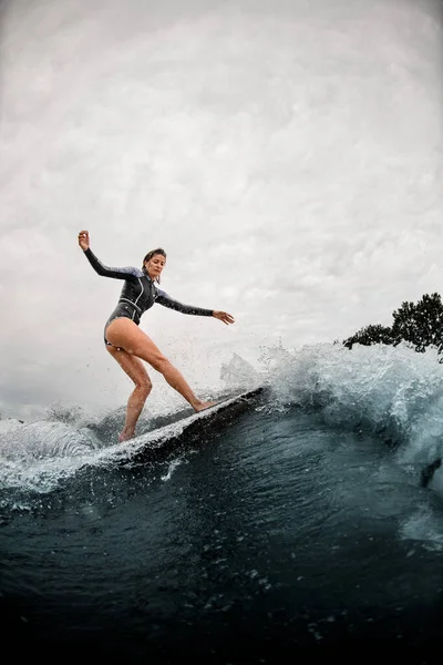 Young woman confidently stands on the wake surf board and rides the wave. — Stock Photo, Image