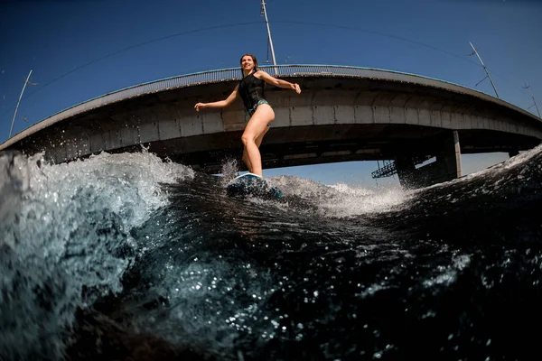 Frau auf Surfbrett reitet die Welle auf dem Hintergrund des Flusses und der Brücke — Stockfoto