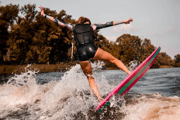 Visão traseira da jovem mulher atraente que fica no wakeboard estilo surf e salta na onda. — Fotografia de Stock