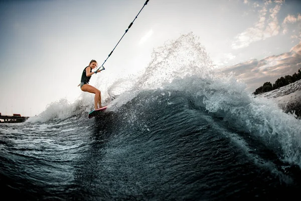 Vista de la mujer joven en wakeboard estilo surf que se aferra a la cuerda y paseos en ola desde el barco —  Fotos de Stock