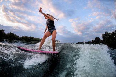 Enerjik genç bir kadın sörf stili Wakeboard 'un üzerinde duruyor ve su sıçratan dalgaların üzerinde dengede duruyor.