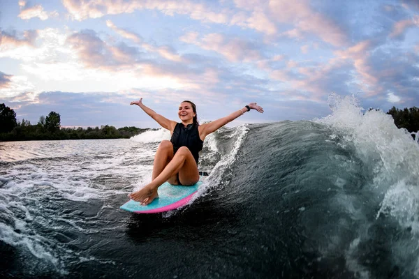 Mujer alegre montar ola mientras está sentado en wakeboard estilo surf con los brazos extendidos —  Fotos de Stock